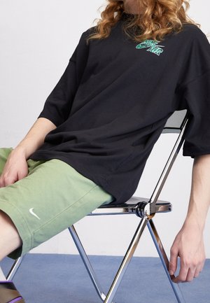 Camiseta negra de gran tamaño con gráfico verde, combinada con pantalones cortos verde claro que presentan un logo blanco de Nike. Modelo sentado en una silla plegable de cromo.