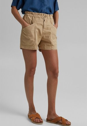 Personne portant un short beige avec des poches avant, une chemise bleue et des sandales marron à brides croisées, debout avec les mains dans les poches.