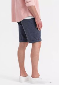 Shorts en tissage bleu marine avec un tissu texturé, des revers roulés et une coupe décontractée, associés à des baskets blanches et une chemise rose pâle.