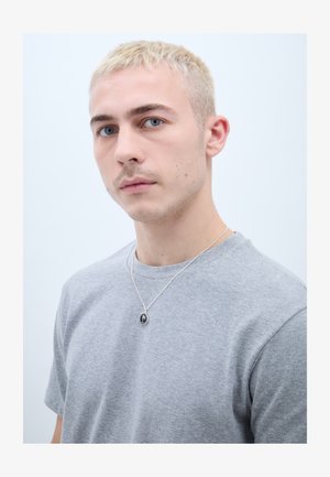 Jaunas vyras su trumpais šviesiais plaukais, dėvintis pilką marškinėlį ir sidabrinį vėrinį su juodu pakabuku, žvelgiantis šiek tiek į šoną.