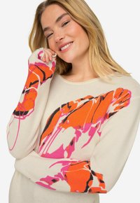 Jasno beżowy sweter z żywym pomarańczowym i różowym wzorem kwiatowym na rękawach. Miękka dzianina, szeroki dekolt i luźny krój.