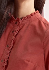 Camicia di velluto a coste rossa con colletto arricciato e chiusura a bottoni. Texture visibile e leggero lucido; presenta una tasca sul petto.