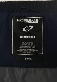 Schwarzfarbiges Pflegeetikett mit weißem Text; identifiziert die Marke "CARISMA," Produkttyp "OBERBEKLEIDUNG," Größe "KLEIN," und Region "NORDAMERIKA."