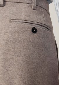 Gros plan d’un pantalon en laine beige avec une poche passepoilée arrière sécurisée par un bouton noir gravé de "FACONNABLE."