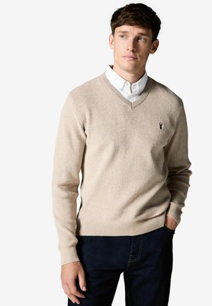 Hellbeige, gestrickter V-Ausschnitt-Pullover aus strukturiertem Material, mit gerippten Bündchen und Saum, kombiniert mit einem weißen Hemd mit Kragen darunter.
