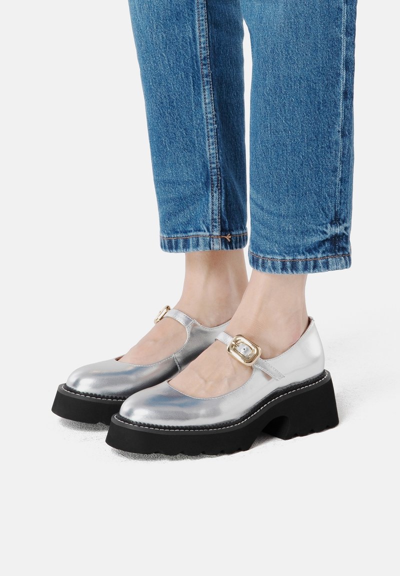 Chaussures Mary Jane métalliques argentées avec un bout arrondi, semelle à plateforme noire et détail de boucle dorée sur la sangle. Portées avec des jeans cropped bleus.
