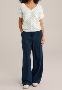 Kobieta ubrana w białą, krótkim rękawem, dzianinową bluzkę na guziki, granatowe spodnie o szerokich nogawkach ze sznurkiem oraz białe trampki z gumowymi podeszwami.