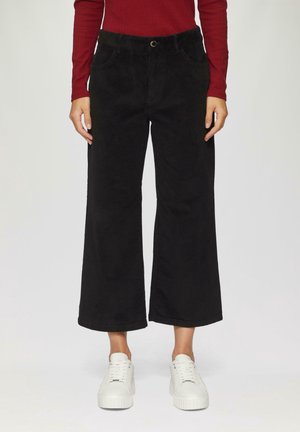 Osoba nosząca czarne szerokie, skrócone spodnie, czerwoną bluzkę z długim rękawem i białe trampki, stojąca na jednolitym tle.