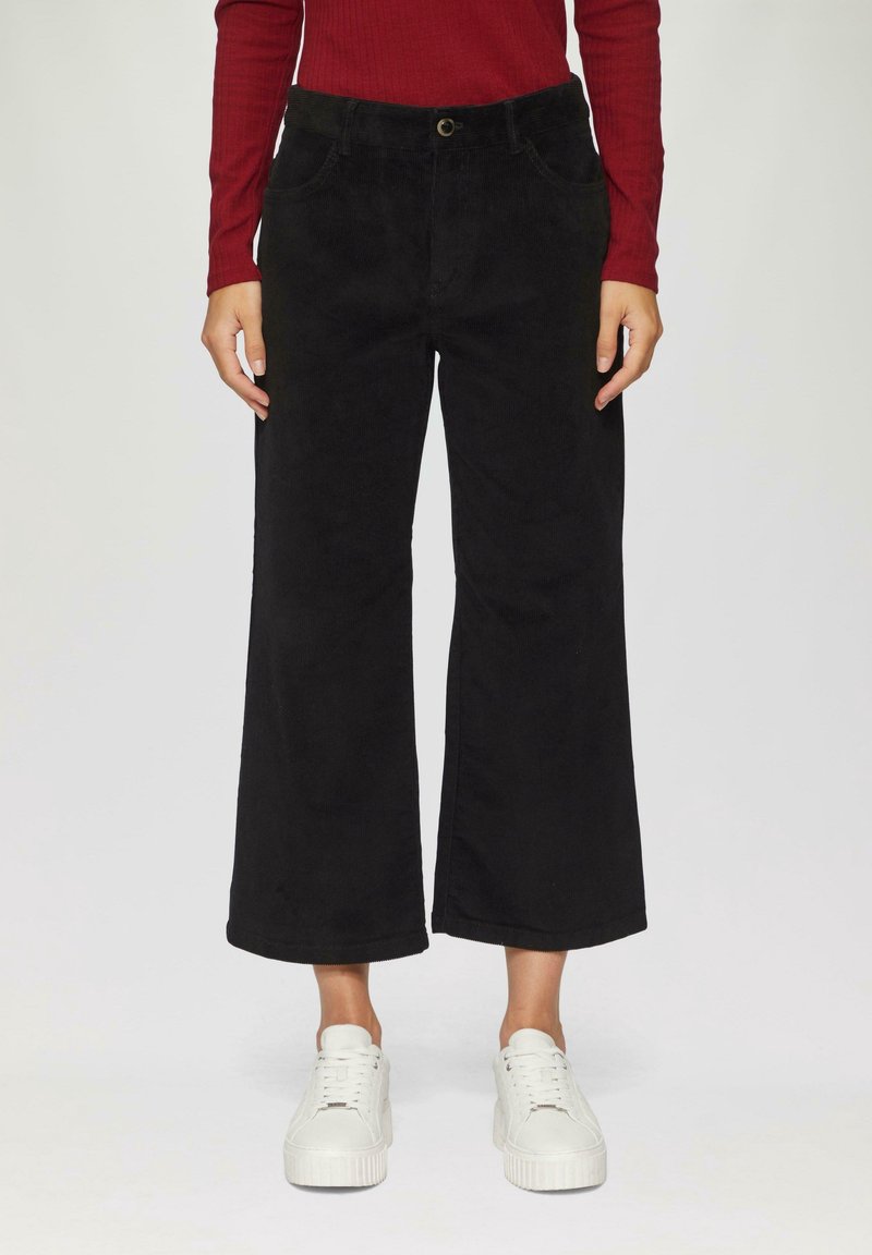 Personne portant un pantalon large noir court, un haut rouge à manches longues et des baskets blanches, debout devant un fond uni.