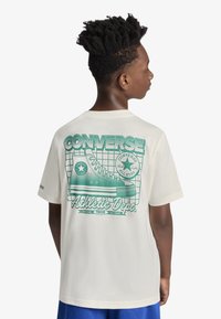 Μπλούζα σε κρεμ απόχρωση με ένα στυλιζαρισμένο γαλάζιο γραφικό ενός αθλητικού παπουτσιού Converse, με το κείμενο «CONVERSE Athletic Dept.» και φόντο με μοτίβο πλέγματος.