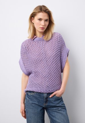 Giovane donna con capelli biondi che indossa un maglione lavorato a maglia lavanda a maniche corte sopra una camicia con colletto e jeans blu, in piedi contro un muro bianco.