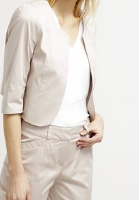 Femme ajustant un blazer beige court et un pantalon taille haute assorti par-dessus un haut blanc sans manches, sur un fond uni.