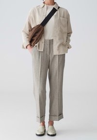 Chemise beige à rayures, haut blanc, pantalon gris texturé avec des bandes latérales, manches retroussées, et mocassins gris clair. Sac bandoulière marron.