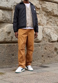 Prošívaná černá bunda přes vzorovanou béžovou vrstvu, doplněná béžovými kalhotami typu cargo a bílými teniskami s tyrkysovými detaily. Pozadí z texturovaného kamene.