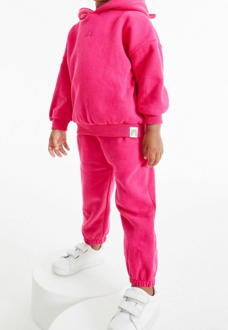 Enfant portant un sweat à capuche rose vif et un pantalon assorti, avec des baskets blanches à scratch, debout sur des plateformes circulaires blanches.
