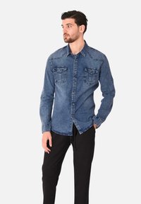 Camicia di jeans blu medio con chiusura a bottoni, due tasche sul petto e maniche lunghe, abbinata a pantaloni neri. Presenta cuciture classiche.