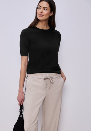 Mujer de cabello largo y oscuro, vestida con una camiseta negra de manga corta y pantalones beige con cordón, sosteniendo un bolso negro, de pie frente a un fondo liso.