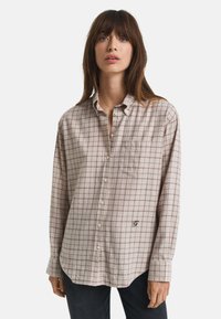 Mulher vestindo uma camisa de botão xadrez bege com um bolso no peito e logo bordado, combinada com calças escuras, em fundo branco.