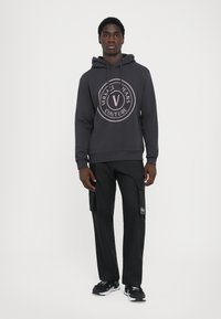 Čierna mikina s kapucňou s oblečeným kruhovým logom "Versace Jeans Couture" v svetloružovej farbe, spárovaná s čiernymi cargo nohavicami a čiernymi teniskami.