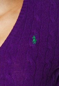 Pull en tricot violet avec un motif en cable et un petit logo vert représentant un cheval et un cavalier sur le côté gauche de la poitrine.