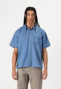 Light blue denim short-sleeve shirt with a button-up front, single chest pocket, and small white logo on the pocket. Worn with light gray pants.
