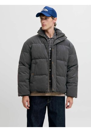 Mladi moški, ki nosi modro kapo, sivo podloženo jakno s pritiskači, karo srajco in temne kavbojke, stoji pred enostavno svetlo ozadje.