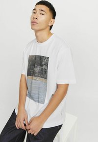 Camiseta blanca de manga corta con una impresión fotográfica de una cancha de baloncesto y texto gráfico. Tejido suave, corte relajado y mínima textura.