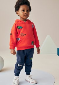 Rød hættetrøje med dyrepatches, stor lomme; kombineret med marineblå sweatpants med en grøn dinosaur og en sjov lomme detalje. Hvide sneakers.