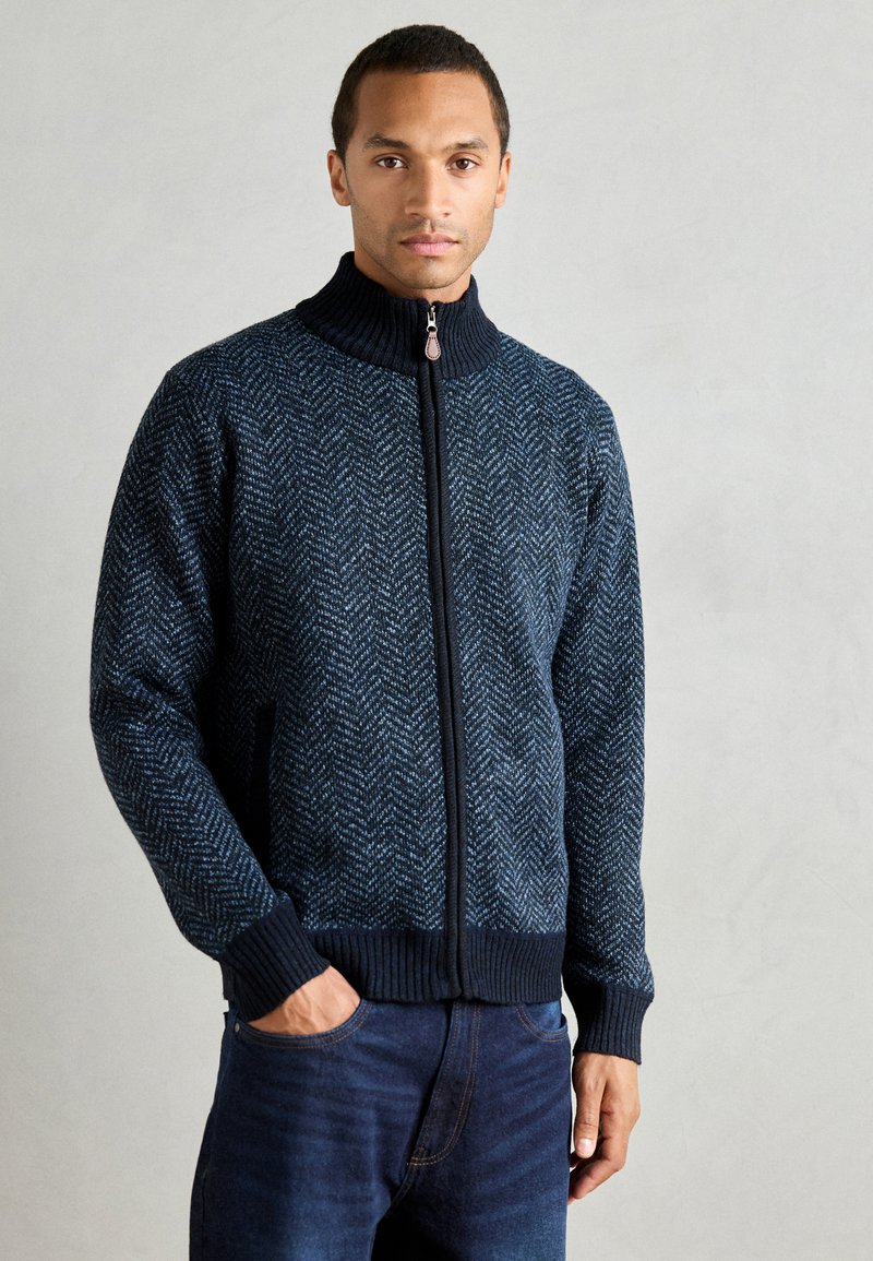 Tricou cu fermoar de culoare bleumarin, cu un model de herringbone, guler înalt și mansete cu nervuri. Fabricat dintr-un material tricotat texturat cu accente întunecate contrastante.