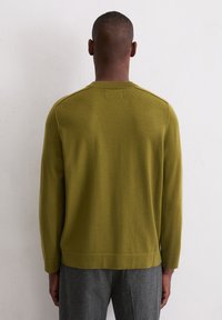 Man in een olijfgroen langhangend sweater en grijze broek, staand met zijn gezicht naar een wit gestructureerde muur en met de handen langs de zij.