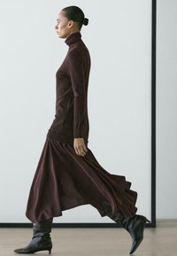 Vínové rolákové šaty s lehce splývavou sukní, vyrobené z měkké látky, doplněné černými kozačkami ke kolenům, zdůrazňující elegantní siluetu.