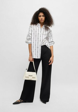 Witte button-up shirt met zwarte verticale patronen, gecombineerd met losse zwarte broek. Model houdt een kleine witte handtas met "HUGO" merk.