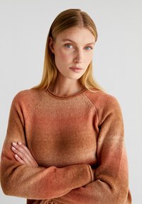 Pull en tricot dégradé dans des teintes d'orange et de marron, avec un col rond, des manches raglan et un tissu doux et texturé.