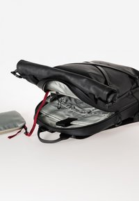 Zaino nero con parte superiore arrotolata, interno grigio e dettagli rossi. Include una pochette argentata staccabile. Materiale liscio e design versatile.