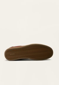 Semelle en caoutchouc avec un motif en zigzag, de couleur marron et d'une finition lisse. Comprend un logo sur le bord latéral.