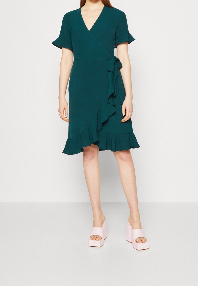 Femme portant une robe portefeuille vert foncé longueur genou avec des bords à volants et des chaussures à plateforme ouvertes roses, debout sur un sol blanc.