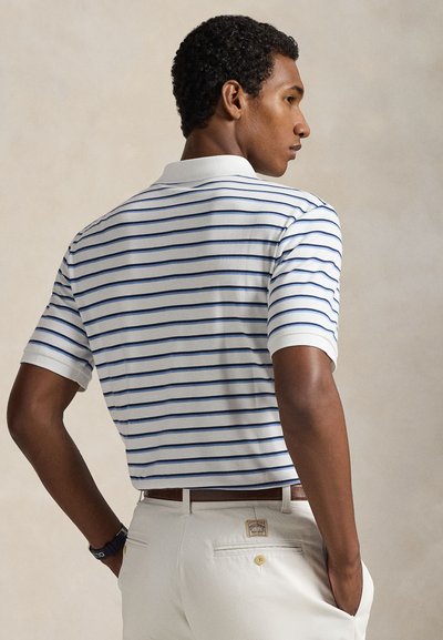 Λευκό πόλο πουκάμισο με μπλε οριζόντιες ρίγες, κοντά μανίκια και κλασικό γιακά. Το μοντέλο φοράει ανοιχτόχρωμο παντελόνι με ζώνη.