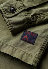 Oliwkowa kurtka z teksturowej tkaniny, z czarnym guzikiem oraz czerwoną i granatową metką z napisem "SUPERDRY UTILITY WEAR."