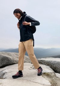Svart vattentät jacka, beige cargo-byxor och svarta vandringsskor med rosa snören. Står på klippor med berg i bakgrunden.