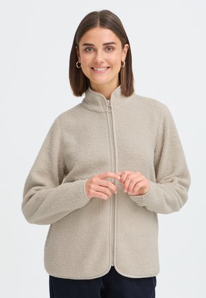 Frau mit schulterlangem braunem Haar, die goldene Creolen trägt und eine beigen Fleecepullover mit Reißverschluss, lächelnd mit verschränkten Händen.