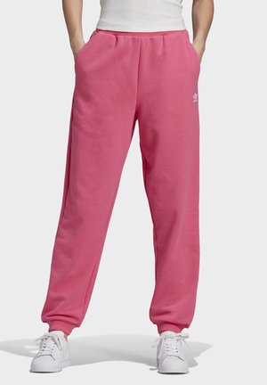 Person iført lyserøde Adidas træningsbukser med lommer, kombineret med hvide sneakers og en hvid top, står med hænderne i lommerne.