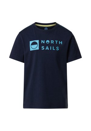 Niño con gorra y camiseta azul marino de North Sails, pantalones cortos turquesa de North Sails, zapatillas blancas, sosteniendo una chaqueta azul marino en una mano.