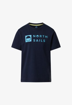 Garçon portant une casquette et un t-shirt North Sails bleu marine, un short North Sails turquoise, des baskets blanches, tenant une veste bleue marine dans une main.
