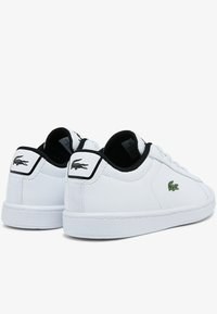 White sneakers with black accents, textured upper, and a green embroidered logo. Features lace-up design and a minimalistic silhouette.