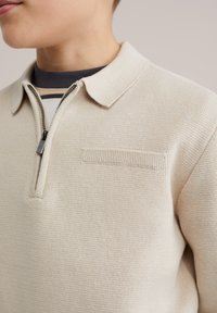 Pull beige à fermeture éclair avec un motif en tricot texturé, doté d'un col et d'une petite poche horizontale sur la poitrine.