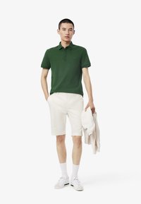 Polo verde, pantaloni corti bianchi, sneaker bianchi e calzini bianchi alla caviglia. Modello che tiene un maglione leggero, in piedi contro uno sfondo semplice.