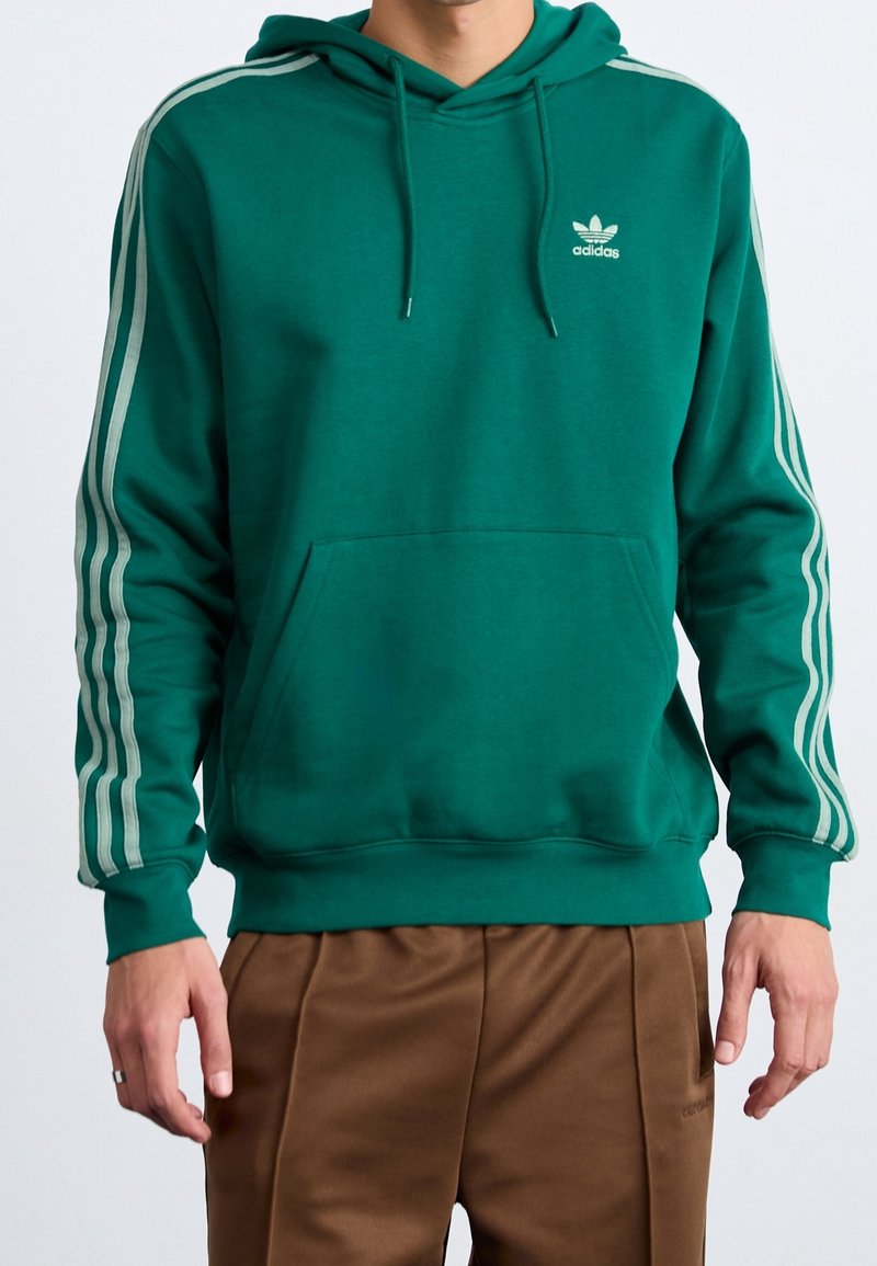 Felpa verde in cotone misto con tasca a marsupio, cappuccio regolabile con coulisse e tre strisce bianche lungo le maniche. Presenta un piccolo logo.