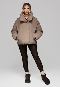 Kurtka puchowa w kolorze taupe z szerokim kołnierzem, z przodem na zamek i kieszeniami bocznymi, połączona z czarnymi legginsami i brązowymi botkami za kostkę.