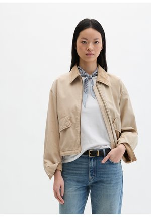 Kobieta o długich czarnych włosach, ubrana w beżową kurtkę, białą koszulę, niebieskie dżinsy oraz wzorzysty jasnoniebieski szalik na szyi.