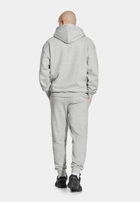 Szary hoodie z dużym kapturem i ściągaczami, w połączeniu z pasującymi dresami. Noszony z czarnymi butami sportowymi, widok z tyłu.